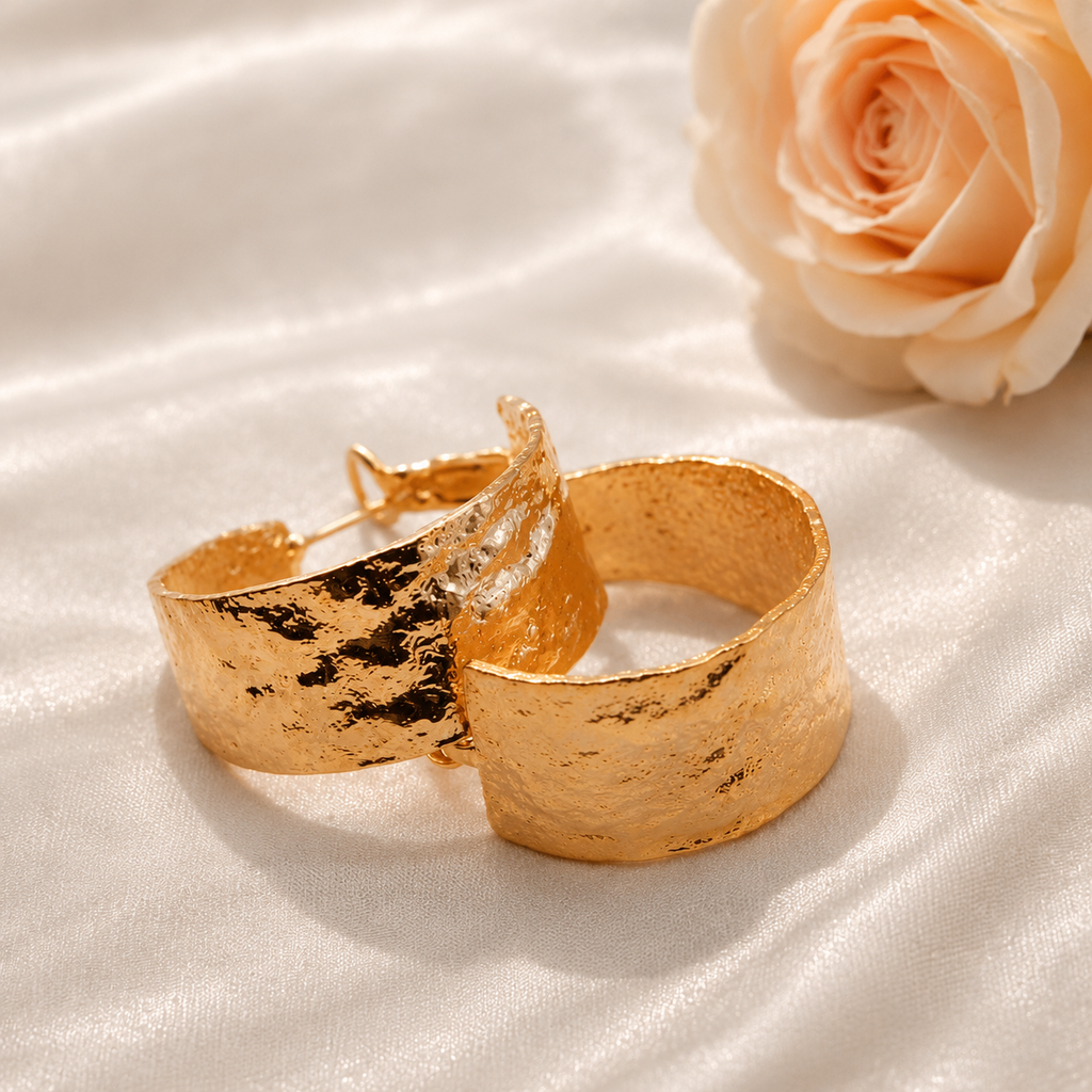 Two textured gold cuff earrings resting on a white satin fabric background with a peach rose in the background.
