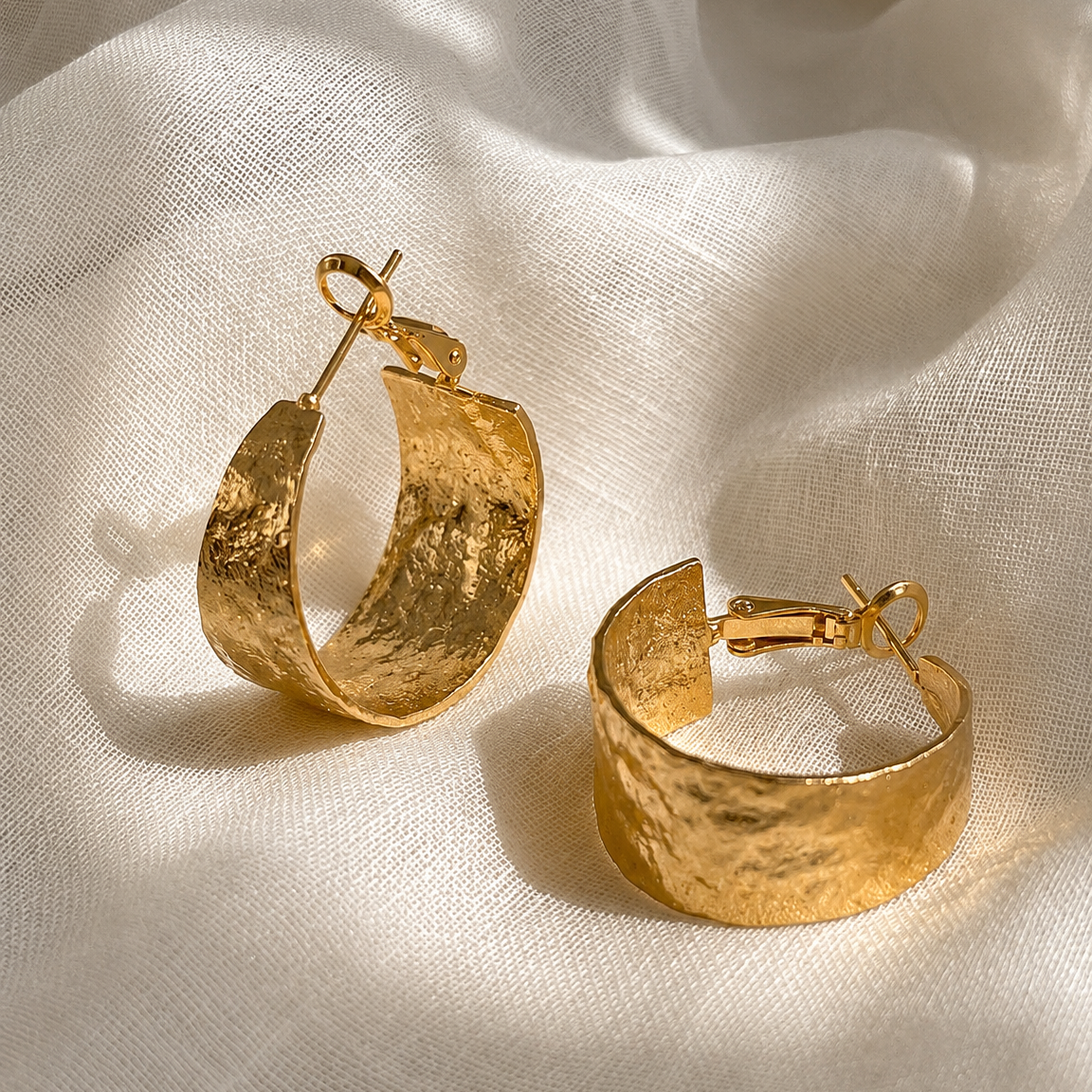 A pair of hammered gold hoop earrings displayed on draped white fabric.