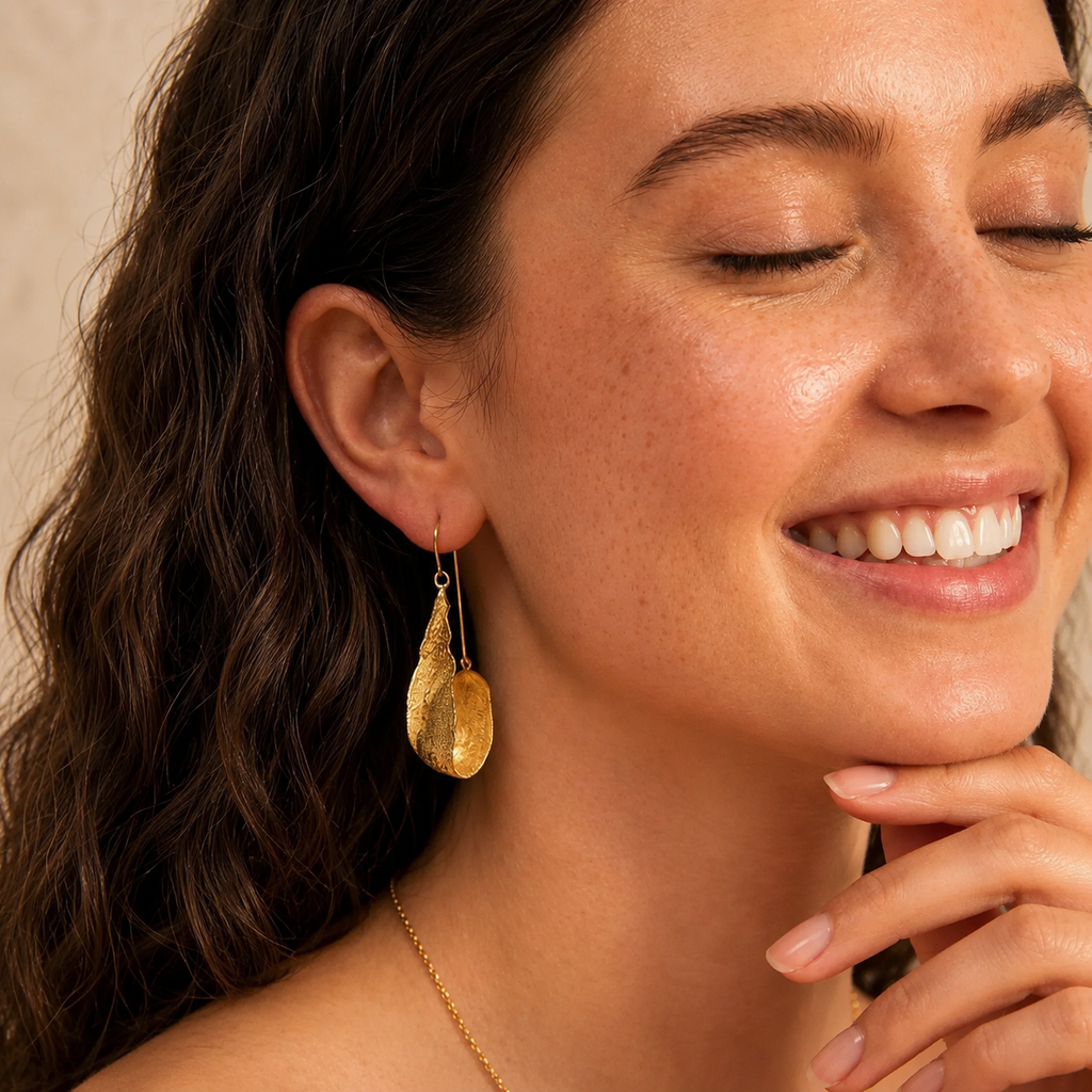 A close-up lifestyle shot of a woman with closed eyes wearing a textured gold teardrop dangle earring and a delicate gold chain necklace, set against a soft beige background.
