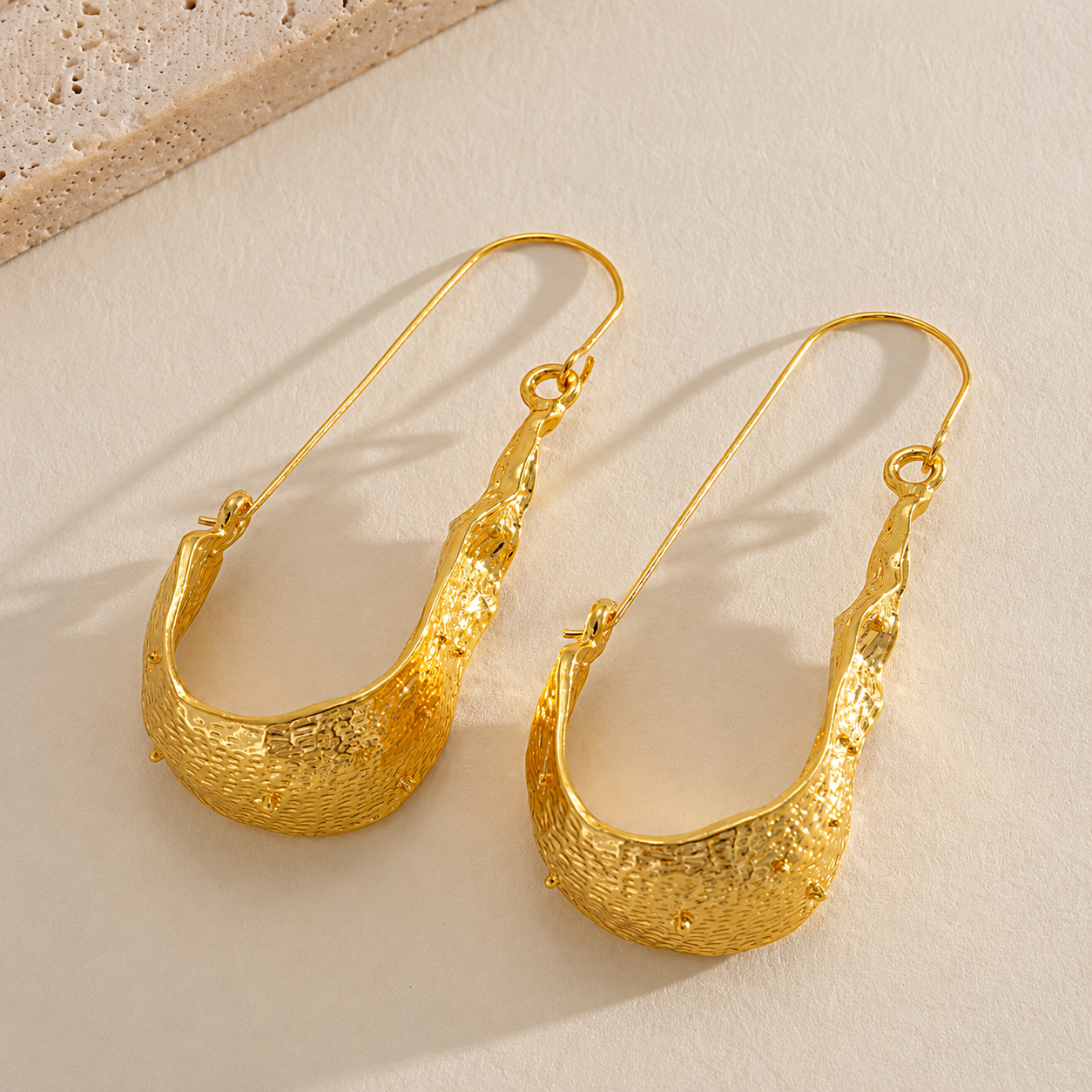 A pair of textured gold hoop earrings with fishhook backings, displayed on a light beige textured surface.