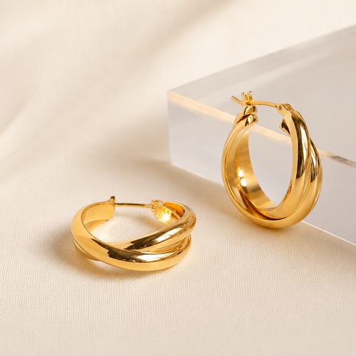 A pair of polished gold hoop earrings, one standing upright on a clear acrylic block and the other lying flat, set against a soft beige fabric background.