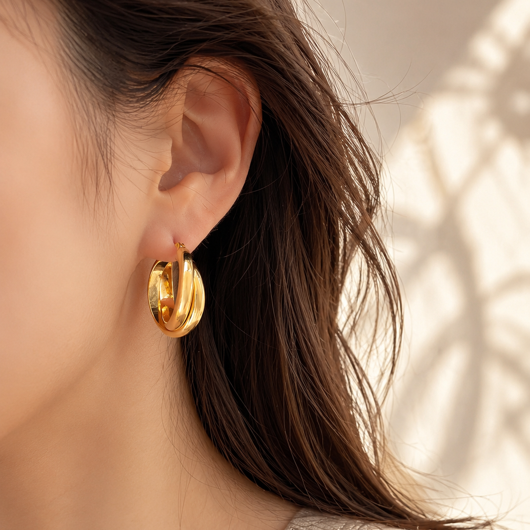 A close-up of a model's ear wearing a polished gold hoop earring, with her hair and a beige ribbed top visible against a softly lit background.