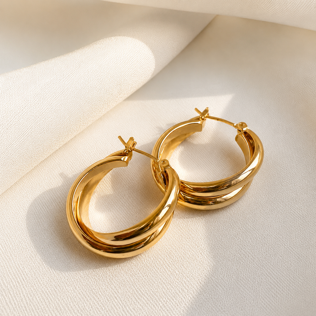 A pair of polished gold hoop earrings with a thick, layered double-band design, resting on a textured cream fabric background with soft shadows.