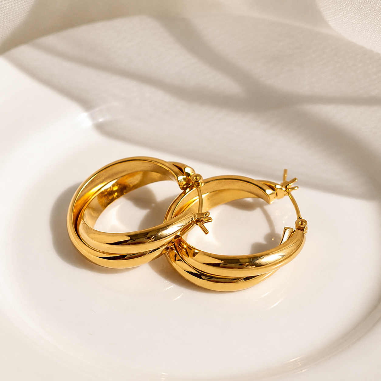 A pair of polished gold hoop earrings resting on a white ceramic plate with soft shadows cast across it.