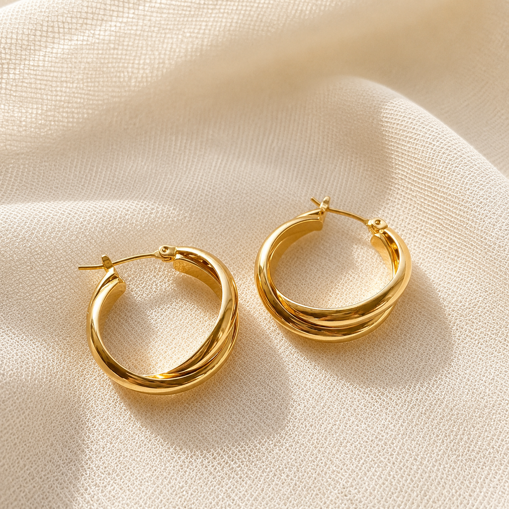 A pair of polished gold hoop earrings with a double-wrap design, displayed on a textured beige fabric background.