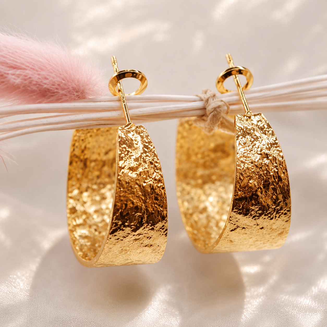 A pair of large textured gold hoop earrings with gold fishhook ear wires, displayed hanging from white strings against a soft, light-colored background with a pink fluffy accent.