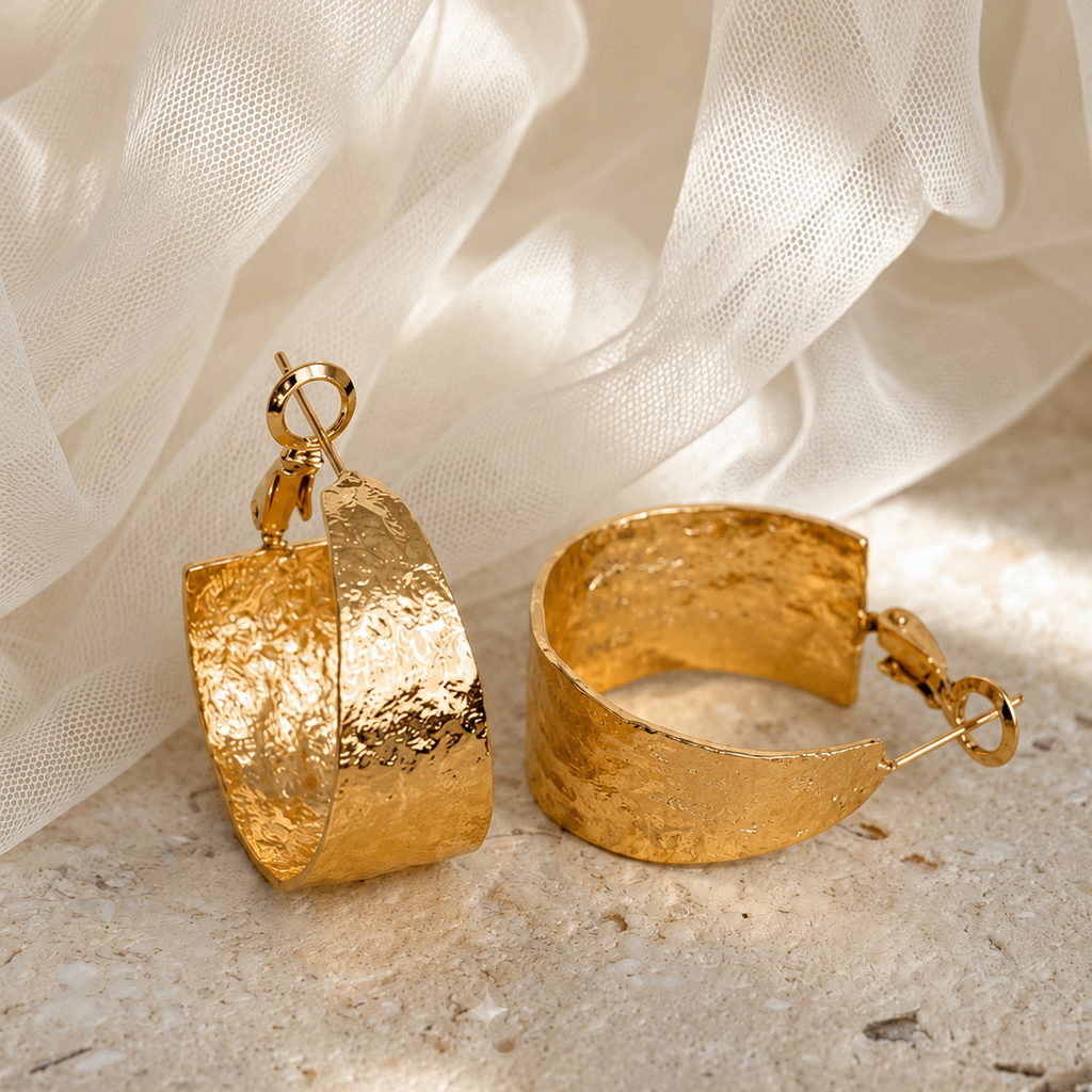 A pair of chunky textured gold hoop earrings with clasp closures, displayed on a light stone surface with white sheer fabric in the background.