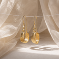 A pair of textured gold drop earrings hanging from a thin wire against a soft beige fabric backdrop with warm sunlight and shadows.