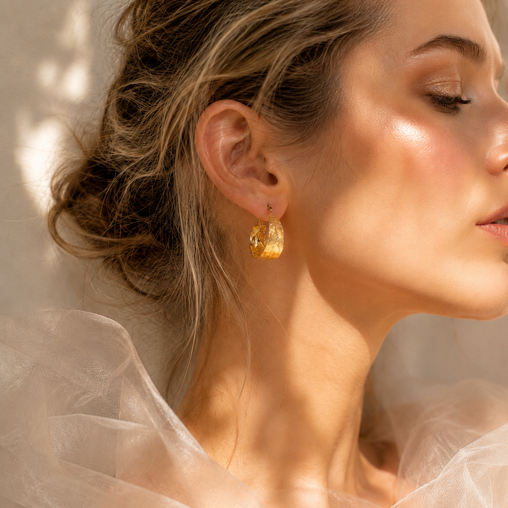 A close-up lifestyle shot of a woman wearing a small, textured gold hoop earring. She is shown in profile with warm sunlight and shadows on her face and neck, wearing a sheer white tulle top.