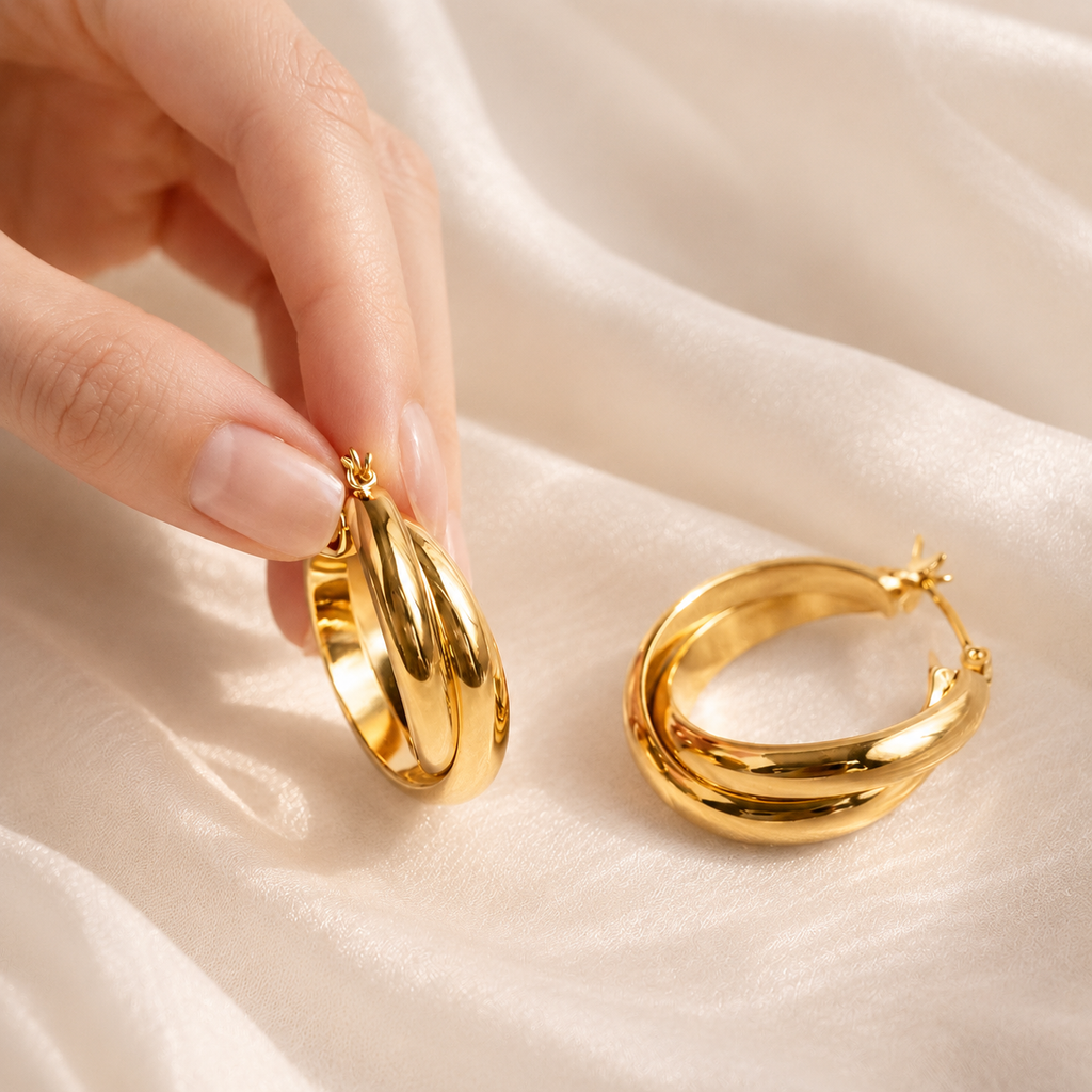 A close-up of a hand holding a pair of polished gold hoop earrings next to another pair resting on draped white satin fabric.